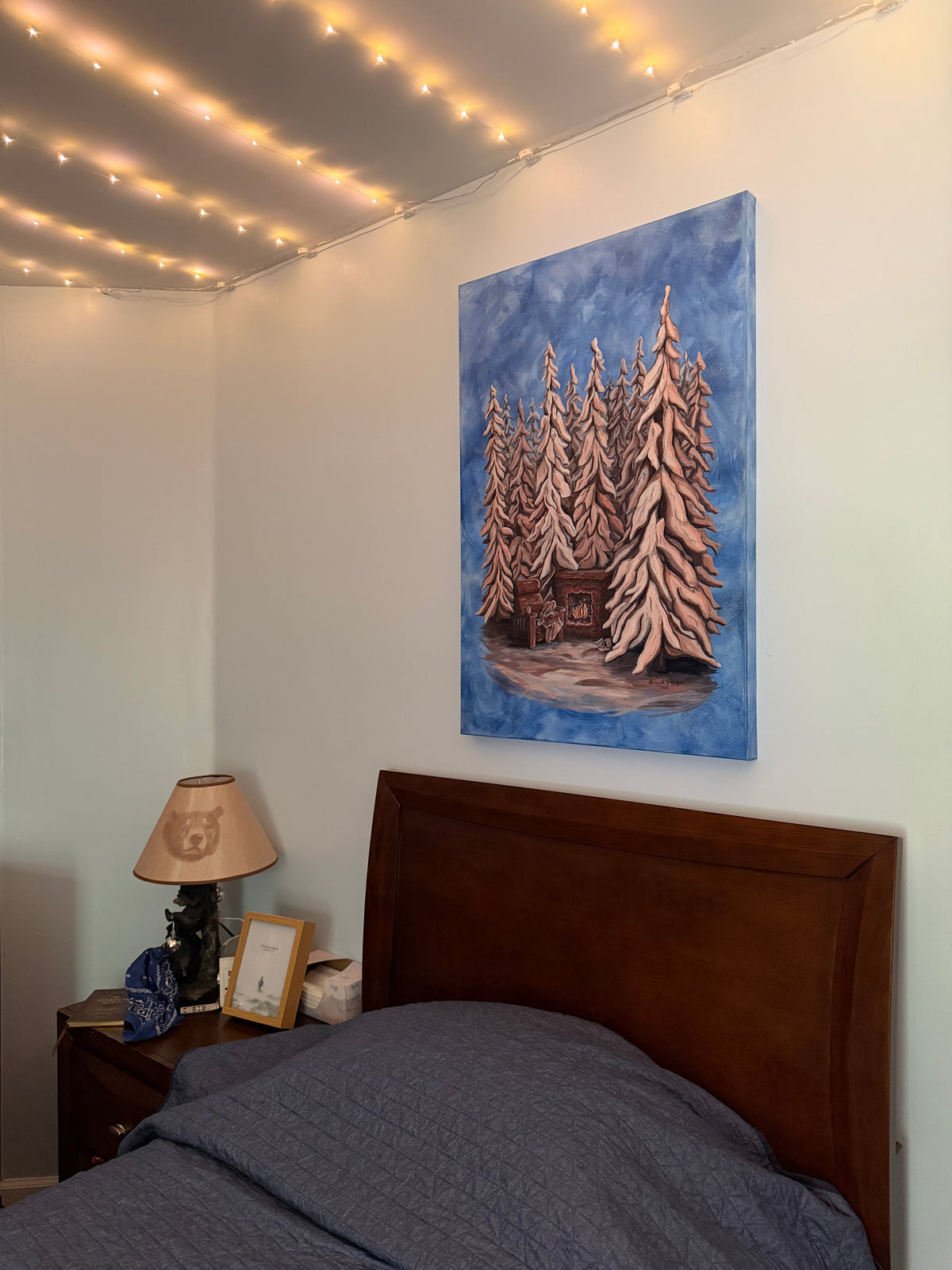 Original painting of a snowy forest scene hanging on a bedroom wall with fairy lights